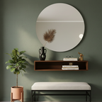 Round mirror above a wooden shelf with decor items against a green wall, next to a plant and bench.