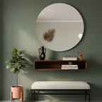 Round mirror above a wooden shelf with decor items against a green wall, next to a plant and bench.