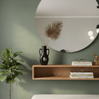 Round mirror on a green wall with a wooden shelf below, containing books, a vase, and decorative items.
